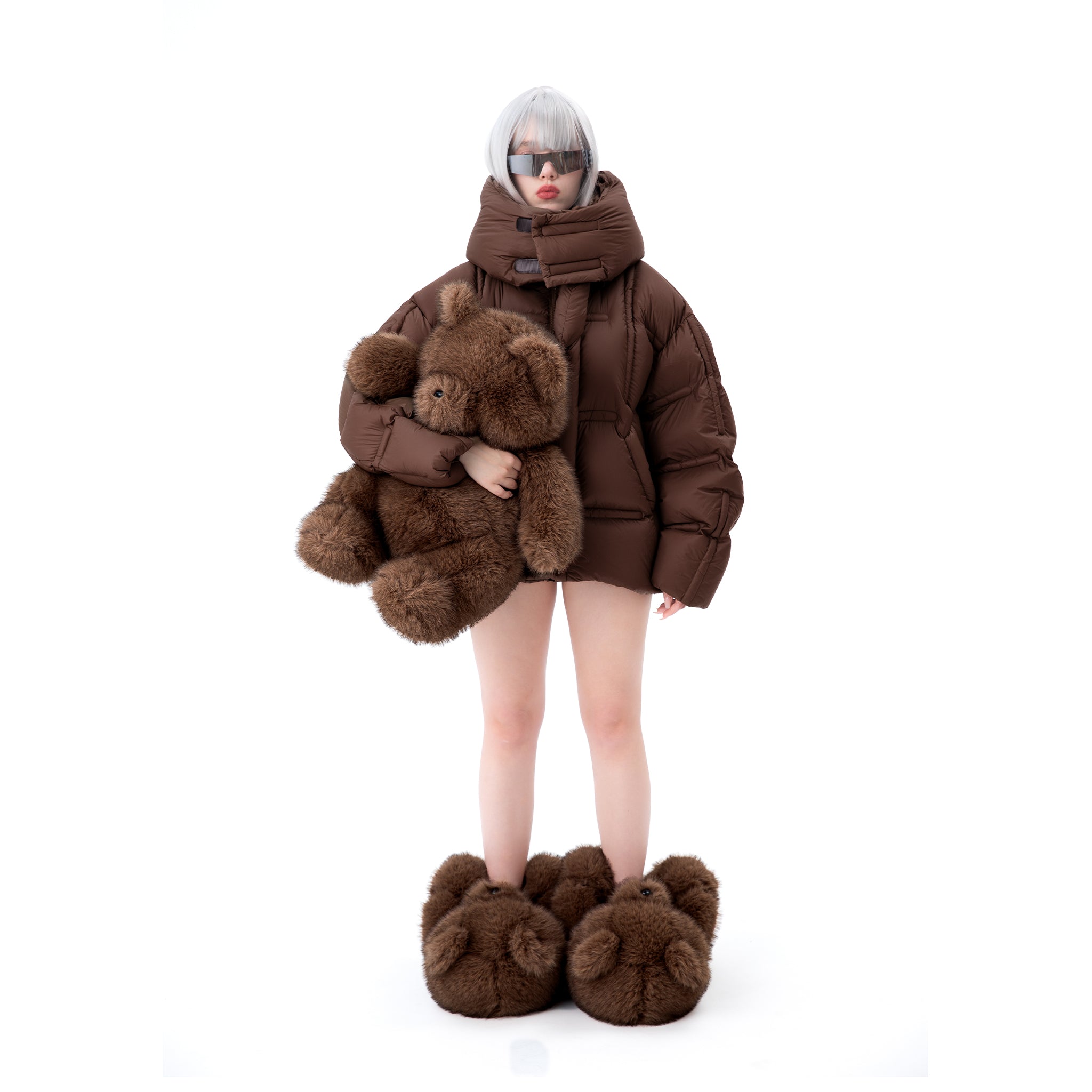 Person girl wearing a brown puffer coat and matching giant teddy bear slippers shoes holding a large brown teddy bear on a white background