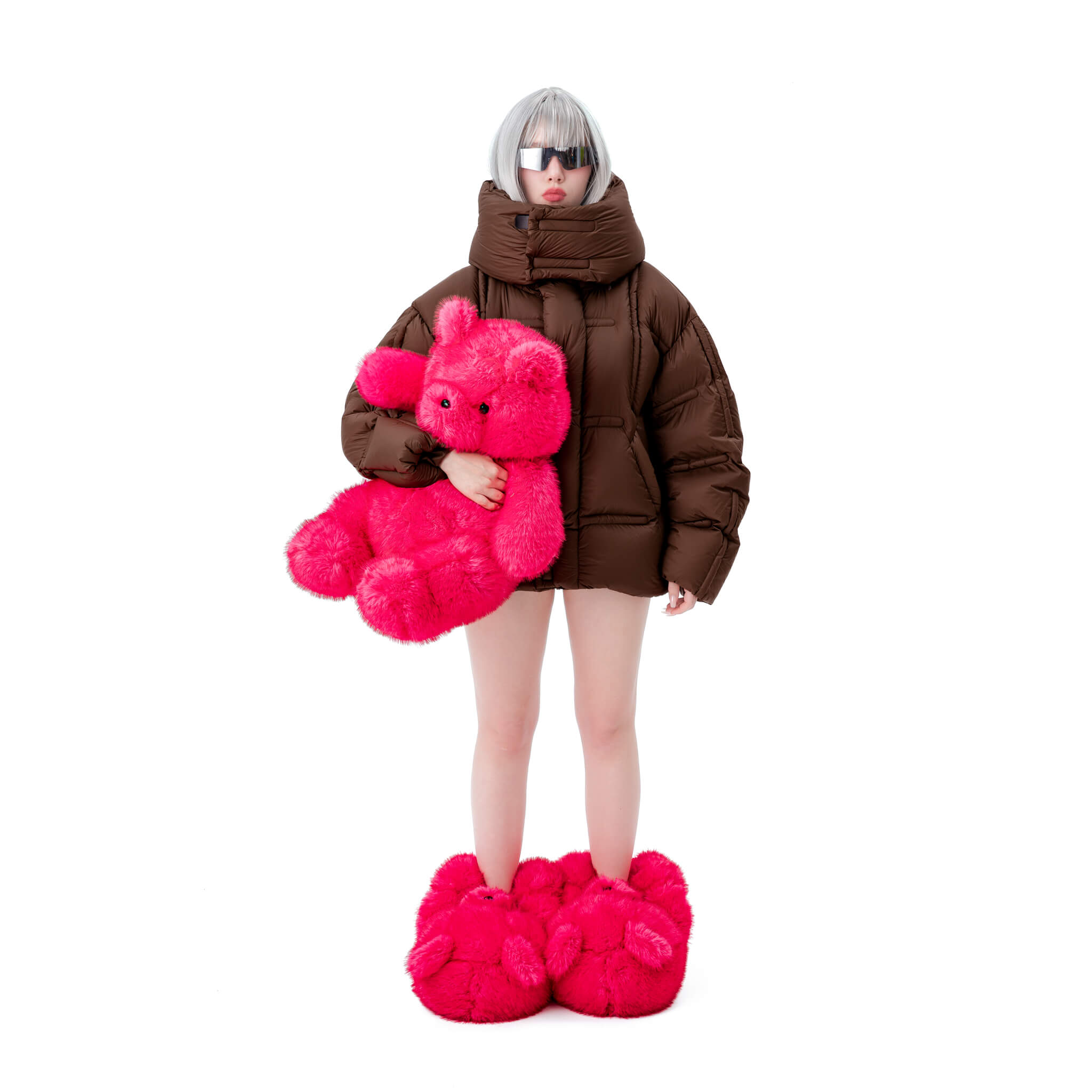 Person girl wearing a brown puffer coat and pink fuzzy big teddy bear slippers shoes holding a pink teddy bear on a white background